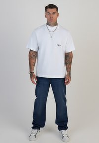 Camiseta de algodón blanca con cuello redondo y mangas cortas, que presenta un pequeño logotipo azul bordado. Combinada con jeans azul oscuro y zapatillas blancas.