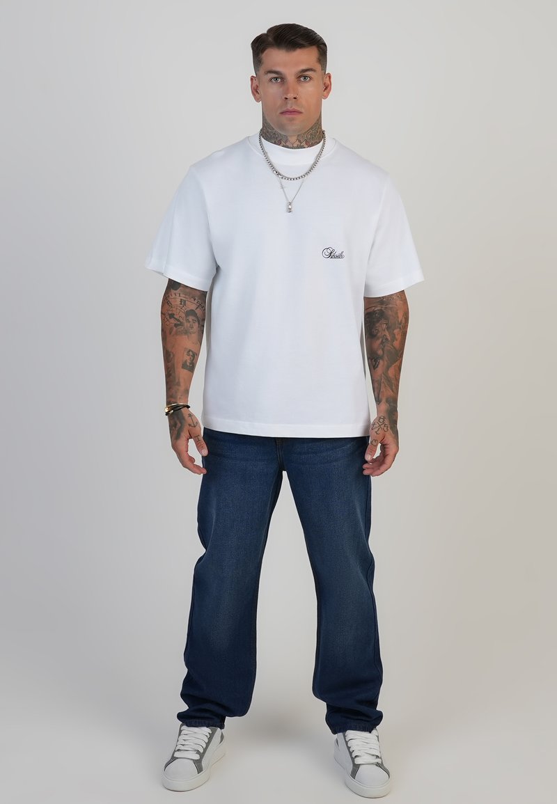 Camiseta de algodón blanca con cuello redondo y mangas cortas, que presenta un pequeño logotipo azul bordado. Combinada con jeans azul oscuro y zapatillas blancas.