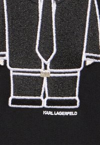 Musta kangas, jossa on valkoinen brodeerattu kuvasto puvusta, solmiosta ja housuista. Teksti "KARL LAGERFELD" on esillä grafiikan alla.