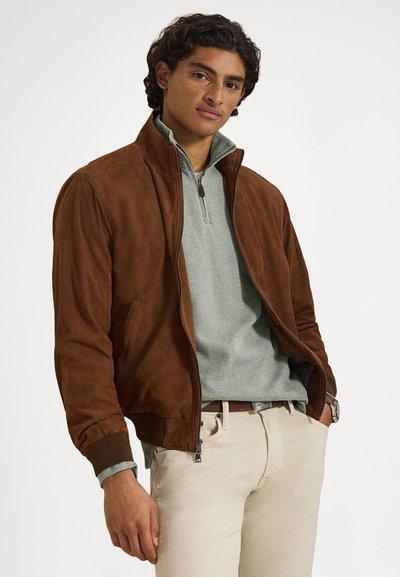 Brown suede bomber jacket with a high collar, zip closure, and ribbed cuffs, paired with a light green mid-layer and beige pants.