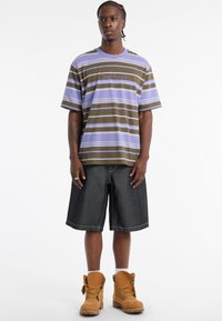 Gestreiftes, kurzärmeliges T-Shirt in Olivgrün und Lavendel, kombiniert mit knielangen dunklen Jeansshorts und braunen Arbeitsstiefeln.
