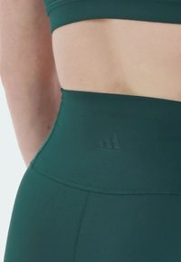 Leggings sportivi verde scuro con vita alta, caratterizzati da un logo in rilievo sottile e una texture di tessuto liscia e elasticizzata.