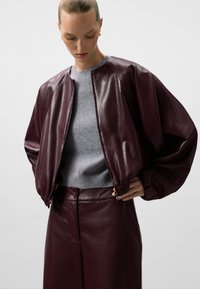 Giacca bomber in pelle bordeaux con maniche voluminose, abbinata a un maglione grigio di maglia e a una gonna in pelle coordinata. Texture liscia e design aderente.