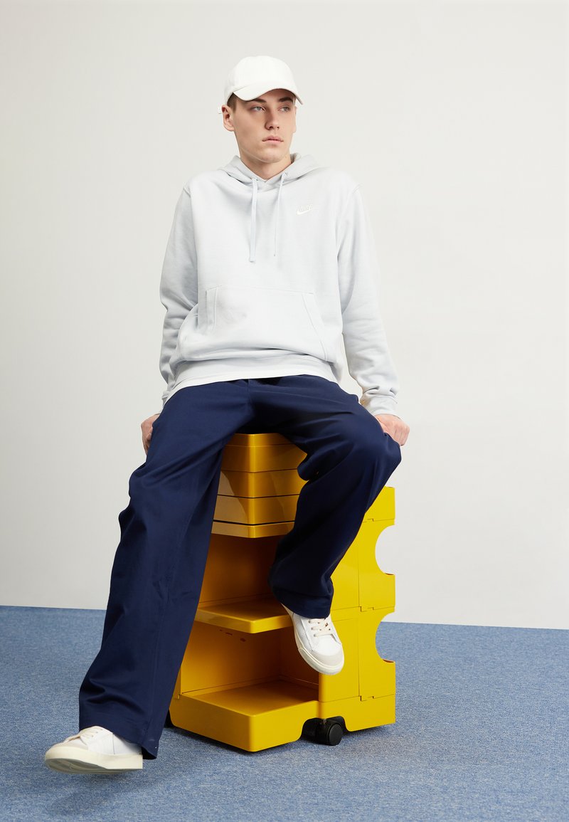 Felpa grigia chiara con tasca frontale e piccolo logo, pantaloni blu navy e scarpe da ginnastica bianche. Modello seduto su un'unità di scaffali modulari gialla.