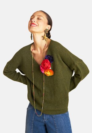 Grüner, lockerer Pullover mit V-Ausschnitt und metallischem Glanz, verziert mit bunten künstlichen Blumen und getragen mit blauen Jeans.
