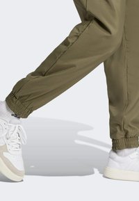 Olijfgroene joggers met taps toelopende boorden, gemaakt van soepele stof, gecombineerd met witte sneakers met beige accenten en met textuur geklede zolen.