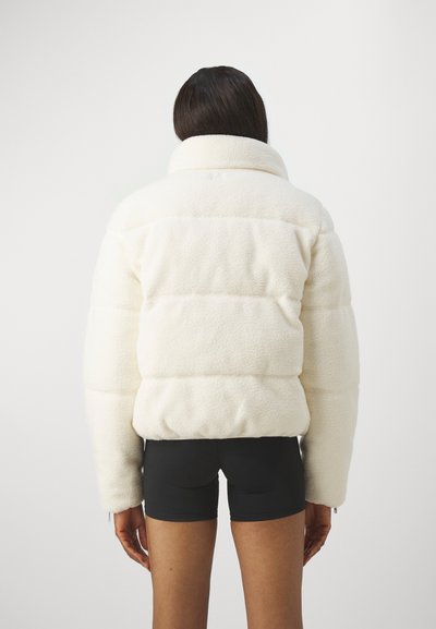 Weiße, flauschige Puffjacke mit hohem Kragen und sichtbaren Nähten, kombiniert mit schwarzen Shorts. Die Textur wirkt weich und plüschig.