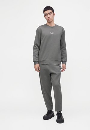 Jeune homme portant un sweat-shirt gris EA7 et un pantalon assorti avec des baskets noires, debout sur un fond blanc.