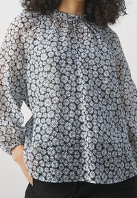 Blouse fleurie en tissu transparent avec des motifs de marguerites noires, bleues et blanches, un col froncé et des manches longues bouffantes.