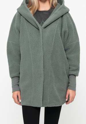 Person trägt eine hellgrüne, übergroße Kapuzen-Fleecejacke mit langen Ärmeln und einem Frontreißverschluss, kombiniert mit schwarzen Hosen.