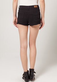 Shorts de mezclilla negra con dobladillo deshilachado, dos bolsillos traseros y una patch de cuero marrón en la cintura. Llevados con botas negras hasta el tobillo.