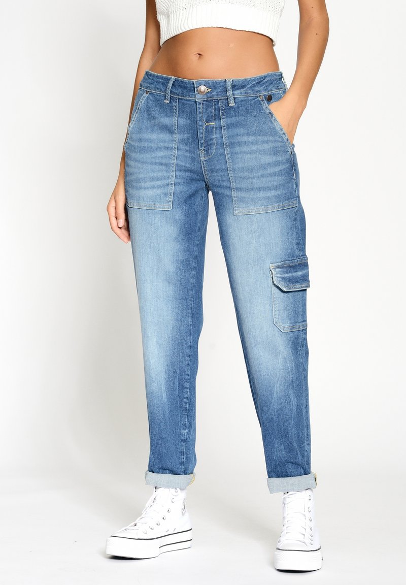 Nainen, joka pukeutunut vaaleansinisiin denimin cargo-housuihin, joissa on etutaskut, sivulla läppätasku, käännetyt lahkeet, valkoiset korolliset lenkkarit ja lyhyt valkoinen paita.