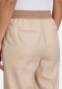 Beżowe spodnie posiadają tkany elastyczny pas, boczne i tylne kieszenie oraz teksturowany materiał z delikatnym połyskiem.