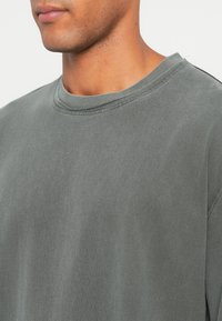 Nahaufnahme einer Person, die ein graues Rundhals-T-Shirt trägt, das den Hals, die Kieferlinie und den oberen Brustbereich zeigt.