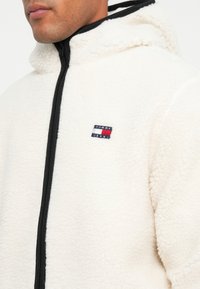 Valkoinen fleece-takki, jossa on musta vetoketju ja huppu. Rinnassa on pieni, brodeerattu "Tommy Jeans" -logo. Pehmeä rakenne.