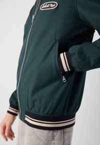 Chaqueta bomber verde hecha de una tela suave, con cierre de cremallera en la parte delantera, puños y dobladillo a rayas, y un logo de parche en el pecho.