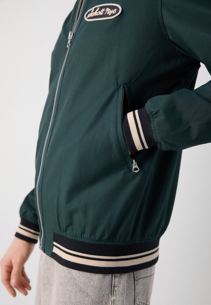 Veste bombardier verte en tissu lisse, avec un zip à l'avant, des poignets et un ourlet rayés, et un logo en patch sur la poitrine.