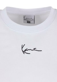 Tricou alb din bumbac, cu guler rotund și semnătură brodată neagră pe piept. Eticheta de interior indică mărimea XS.
