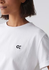 T-shirt bianca in cotone a girocollo con un piccolo logo ricamato nero sul lato sinistro del petto. Maniche corte con orli arrotolati.