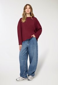 Felpa oversized bordeaux con maniche lunghe, abbinata a jeans di denim blu ampi, e scarpe sportive argento. Tessuto dalla texture liscia.