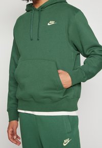 Person som bär en grön Nike-hoodie och matchande sweatpants med krämfärgade logotyper, hand i huppjackans ficka, vit skjorta synlig under.