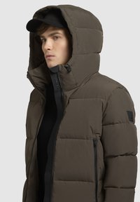 Puffer jacket in dark olive green, featuring a high collar, large hood, front zipper, and black hardware accents. Textured matte finish.