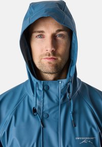 Blå vattentät jacka med huva, med zip-fram, tryckknappar, dragsnören och logotypen på nedre högra sidan.