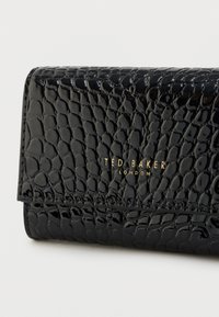 Czarny portfel z lakierowanej skóry z fakturą w kształcie krokodyla oraz złotym logo "Ted Baker London". Wyposażony w zamknięcie na magnetyczną klapkę.