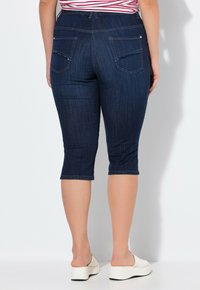 Jeans capri de color azul oscuro, hechos de denim, con una textura suave, cinco bolsillos y detalles deshilachados en los bolsillos traseros. Largo recortado por encima del tobillo.