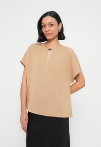 Μπεζ μπλούζα με κοντό μανίκι και άνοιγμα στο λαιμό, με λεία υφή, άνετη εφαρμογή και διακριτική ετικέτα μάρκας κοντά στη λαιμόκοψη. Φορεμένη πάνω από μαύρη φούστα.