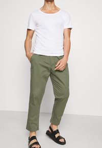 Man in een witte T-shirt met korte mouwen, olijfgroene cropped broek en zwarte open teen sandalen, staand met de handen bij de zakken op een effen achtergrond.