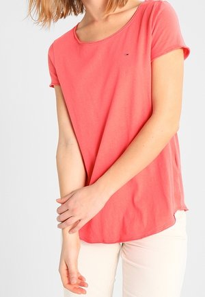 Nainen, jolla on päällään korallinvärinen lyhythihainen t-paita ja vaaleanbeiget housut, ristii käsivartensa ranteiden kohdalla yksinkertaista taustaa vasten.