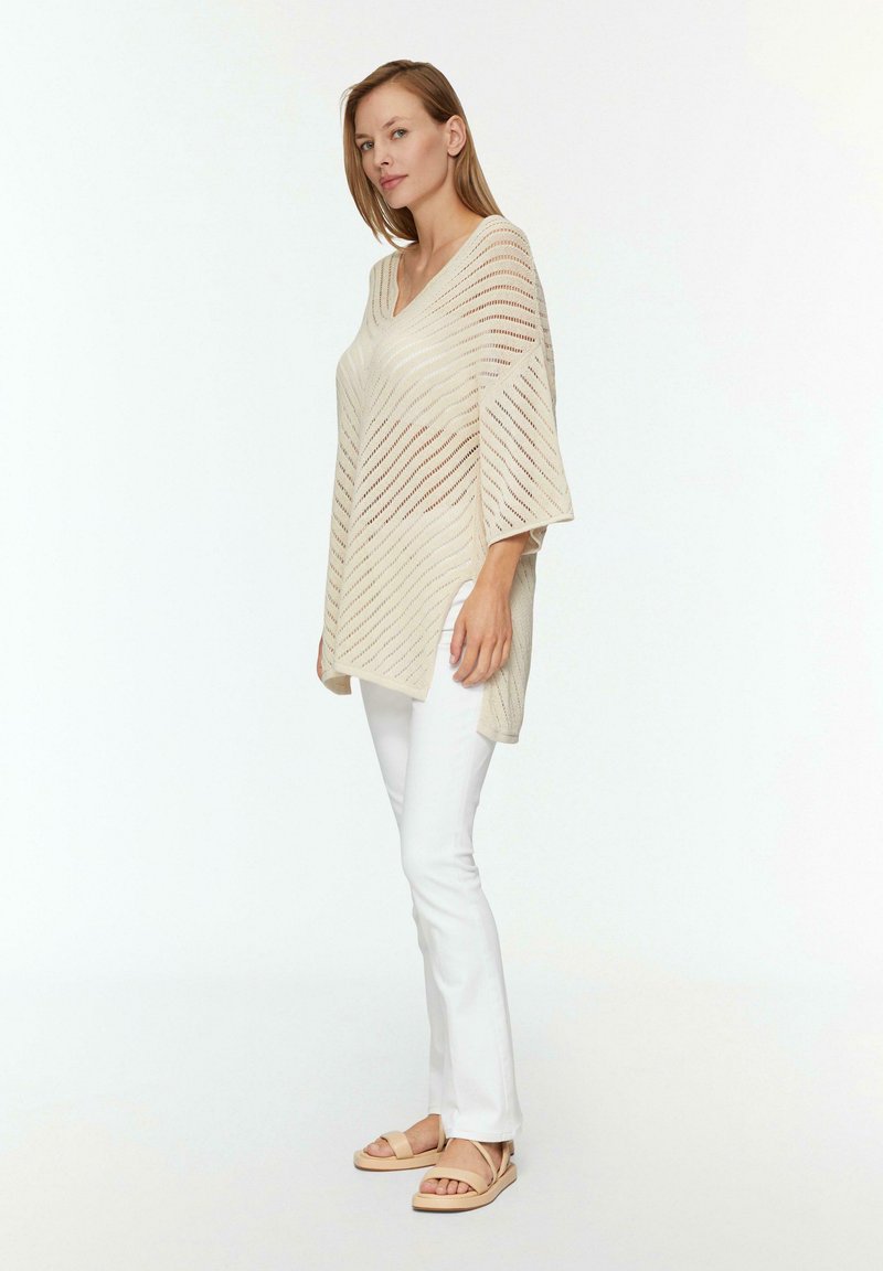 Beige gebreide poncho met een V-hals, met diagonale streepjespatronen. Gecombineerd met witte slim-fit broeken en neutrale sandalen.