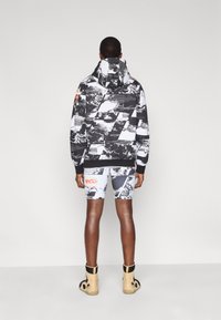 Φούτερ με κουκούλα σε μαύρο και λευκό με γραφικό σχέδιο, σετ με σορτς που φέρουν παρόμοιο μοτίβο. Το μοντέλο φοράει μπεζ παπούτσια τύπου high-top.