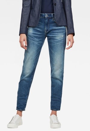 Personne portant un jean bleu délavé, un blazer sombre, un haut bleu marine et des baskets blanches, debout les bras détendus le long du corps.
