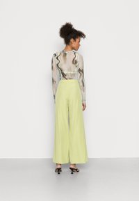 Blusa transparente de mangas longas com padrões abstratos em castanho e bege; calças largas de cintura alta em verde-claro; sandálias de salto pretas.
