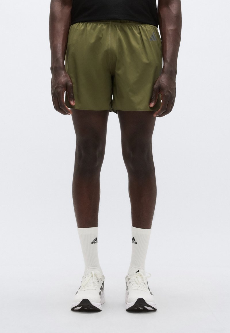 Πράσινο αθλητικό σορτς με ελαστική μέση, με αντίθετα λογότυπα, φορεμένο με λευκές κάλτσες πλήρους μήκους και παπούτσια τρεξίματος με μαύρες λεπτομέρειες.