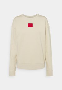 Beżowy sweatshirt z okrągłym dekoltem, długimi rękawami i ściągaczami. Ma czerwony prostokątny haft z logo "HUGO" na froncie.