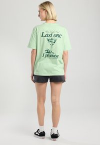 Mint green T-shirt featuring a printed graphic of a cocktail glass and text on the back. Paired with black shorts and patterned sneakers.
