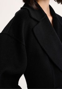 Czarny wełniany płaszcz z wciętym kołnierzem, oversize'owymi rękawami i miękkim, fakturowanym wykończeniem. Proste linie z minimalną ilością detali.