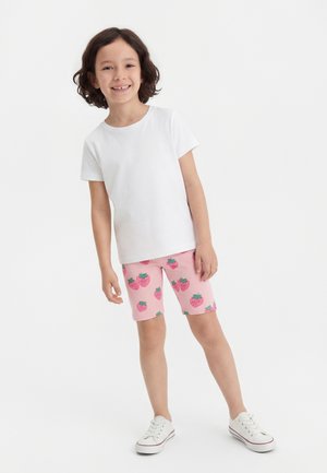 Smiling child standing, wearing a plain white t-shirt, pink shorts with strawberry prints, and white sneakers against a plain white background.