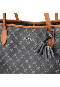 Bolso tote de cuero gris con un patrón blanco repetido, detalles en tono marrón y un logo dorado. Presenta detalles de borlas adjuntas al mango.