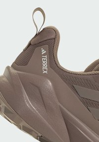 Braunes Sportschuh mit strukturiertem Obermaterial, verstärktem Fersenbereich und einem auffälligen geometrischen Sohlenmuster. Markenlogo an der Seite sichtbar.