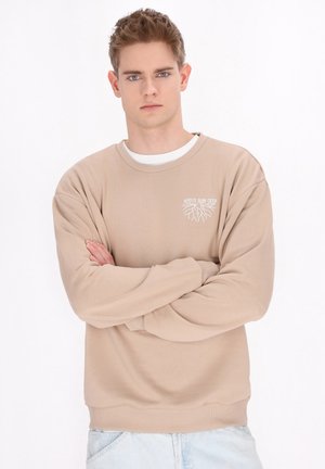 Beige sweatshirt met lange mouwen, geribbelde manchetten en ronde hals. Kenmerkend is de witte gedrukte grafiek op de borst. Gestyled met lichtblauwe denim jeans.