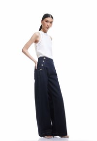 Valkoinen hihaton paita, jossa on pitsiyksityiskohtia, yhdistettynä korkeavyötäröisiin, väljiin tummansinisempiin farkkuihin, joissa on kultanappiyksityiskohtia ja sivutaskut.