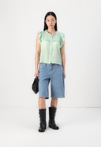Blusa verde sem mangas com gola fruncida, combinada com calções de jeans azuis até ao joelho e botas pretas até ao joelho. Mala preta na mão.