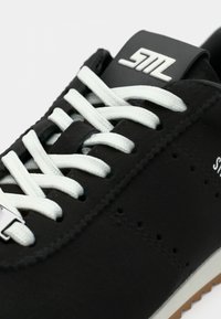 Μαύρο αθλητικό παπούτσι με λευκά κορδόνια, λαστιχένια σόλα και λεία υφή. Διαθέτει λογότυπο στη γλώσσα και μικρές οπές αερισμού στο πλάι.