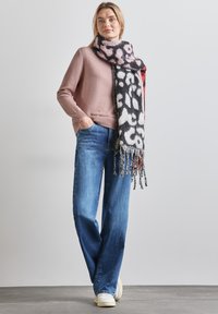 Maglione rosa chiaro, jeans a gamba larga blu e una sciarpa a contrasto bianco e nero con frange. In piedi su un pavimento grigio con uno sfondo neutro.
