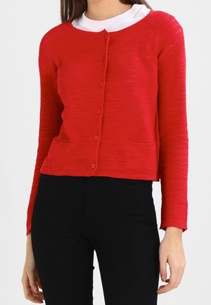 Gilet en maille côtelée rouge, à manches longues, avec six boutons, encolure ronde et deux poches avant, porté sur une chemise à col blanche en dessous.
