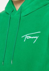Felpa verde in pile con cappuccio regolabile. Presenta un logo ricamato bianco "Tommy" sul davanti. Tessuto morbido.